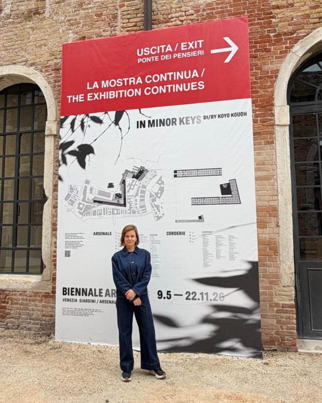 Jess, a white woman, stands in front of a large white poster that's advertising the Venice Biennale